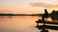 Man in silhouette fishing from wooden dock at golden sunset on calm lake, peaceful outdoor recreation and leisure activity for relaxation and nature themes.