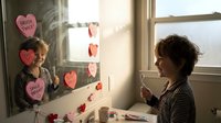 Young child with curly hair creating Valentine's Day heart decorations on window, crafting holiday cards with pink and red paper hearts at home.