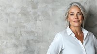 Confident mature woman with silver gray hair wearing white shirt standing against textured concrete wall background, natural beauty and aging gracefully concept.