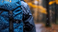 Wet blue backpack with water drops in rainy autumn weather, close-up view against blurred orange fall foliage background outdoors.