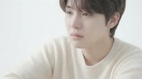 Young East Asian man with tear on cheek wearing cream sweater, looking away with sad expression against white background.