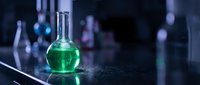 Green chemical solution in round bottom flask on laboratory bench with blurred glassware background for scientific research and experimentation.