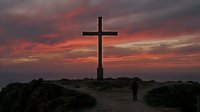 Silhouette of wooden cross against dramatic sunset sky with person walking on path. Religious symbol for faith, spirituality and worship concepts.
