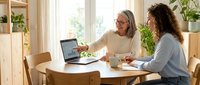 Two women collaborating at home office desk with laptop computer discussing business project in bright modern interior with plants and natural lighting.