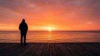 Silhouetted figure standing on wooden pier watching golden sunset over calm ocean water, contemplative mood for travel and lifestyle content.