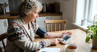 Senior Caucasian woman measuring blood pressure at home kitchen table with digital monitor device for health monitoring and self-care routine.