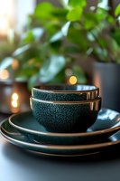 Elegant dark green ceramic bowls and plates with textured glaze finish stacked on table, soft bokeh lights and green plant leaves in background. Modern tableware styling.