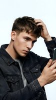 Young Caucasian male teenager wearing dark denim jacket, touching hair with hand while looking at smartphone screen against white background for youth lifestyle and technology concepts.