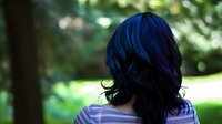 Young Asian woman with vibrant blue-black hair shown from behind in natural outdoor setting with soft bokeh background and sunlit foliage. Wearing casual striped top.