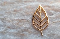 Golden leaf-shaped brooch with delicate veining pattern on textured gray marble background, minimalist jewelry design showcasing nature-inspired elegance.