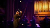 Orchestra conductor leading classical music performance in elegant concert hall with pipe organ and musicians in background during evening show.