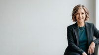 Mature woman with shoulder-length blonde hair, Caucasian ethnicity, wearing professional black blazer and teal top, smiling confidently against white background. Corporate portrait for business, leadership, executive roles.