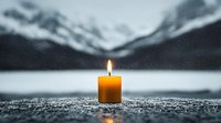 Single burning yellow candle against blurred winter mountain landscape background with snow and lake, creating atmospheric moody scene with warm candlelight glow.