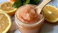 Pink grapefruit and lemon sugar body scrub in glass jar with wooden spoon, fresh citrus slices and mint leaves on blue background for spa treatment.