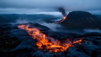 Dramatic volcanic eruption with glowing lava flows cascading down mountainside at dusk, creating striking contrast against dark landscape and water below.