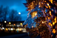 Illuminated wire mesh sculpture of human face profile against dark blue night sky with bokeh lights and full moon, creating mystical atmosphere.