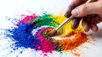 Unrecognizable hand holding brush applying vibrant colored powder pigments arranged in rainbow pattern on white surface for art projects and creative design work.