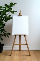 Wooden easel with blank white canvas against neutral wall, green plant in corner. Minimalist art studio setup on hardwood floor for painting or presentation.