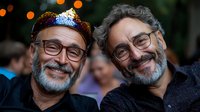Two mature bearded men with glasses smiling warmly at camera, wearing casual attire. One has festive crown. Natural bokeh lights in background create joyful atmosphere.