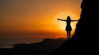 Woman silhouette with arms outstretched on cliff edge during golden sunset over ocean. Freedom, achievement, and spiritual connection with nature concept.