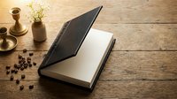 Open leather notebook with blank pages on rustic wooden table with coffee beans, brass candlestick and small plant pot creating cozy writing atmosphere.