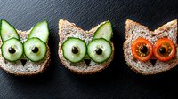 Playful cat-shaped sandwiches decorated with cucumber and tomato slices as eyes, creating fun food art on dark background. Creative kids lunch concept.