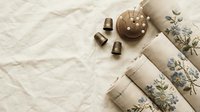 Vintage sewing supplies with floral embroidered linen fabric, wooden buttons and brass thimbles on neutral textile background for crafting projects.