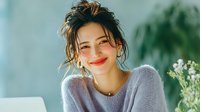 Happy young Asian woman with messy bun hairstyle smiling while working on laptop at home office desk with flowers for lifestyle and remote work content.