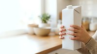 Woman holding white milk carton in bright kitchen with wooden countertop and blurred background, suitable for dairy, beverage, and food packaging concepts.
