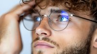 Young Caucasian man with curly hair and glasses touching head in thoughtful pose, professional headshot for business and lifestyle concepts.