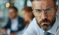 Serious bearded businessman in glasses looking intensely at camera during corporate meeting, with blurred colleagues in background. Professional workplace portrait.