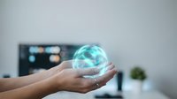 Hands holding glowing holographic sphere in modern office environment with blurred computer screen background for futuristic technology concepts.