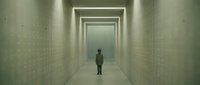 Man standing alone in long narrow corridor with glowing walls and bright light ahead, conveying isolation, uncertainty, and journey through institutional or futuristic space.