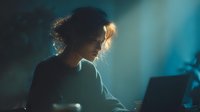 Young woman with curly hair working late at computer in moody blue lighting. Night shift programmer or designer focused on screen in dark office atmosphere.