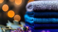 Stack of folded knitted sweaters in blue and gray shades with blurred golden bokeh lights and decorative elements in foreground, cozy winter fashion concept.