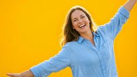 Mature woman with long blonde hair, Caucasian ethnicity, wearing light blue button-up shirt, smiling with arms outstretched in celebratory pose against bright yellow background for lifestyle and wellness content.