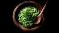 Creamy green pea risotto served in rustic wooden bowl with fresh herbs and wooden spoon, dramatic dark food photography with moody lighting.