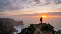 Silhouette of hiker standing on rocky cliff overlooking ocean at sunset, golden hour landscape with dramatic sky and coastal cliffs for travel and adventure themes.