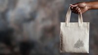 African American hand holding natural cotton eco bag with minimalist tree design against blurred dark background, demonstrating sustainable shopping choice.
