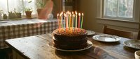 Chocolate birthday cake with colorful lit candles on rustic wooden table in cozy home kitchen with natural light.