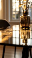 Amber glass vase with dried flowers on glossy black table surface reflecting warm sunlight through window, creating cozy atmospheric home interior with soft bokeh effect.