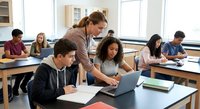 Female teacher helping diverse students with laptop in modern classroom during collaborative learning session.