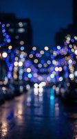 Blurred city street at night with colorful bokeh lights creating dreamy urban atmosphere. Wet pavement reflects illuminated buildings and traffic.