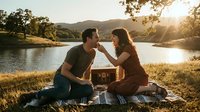 Couple sharing food during romantic lakeside picnic at golden hour with mountains and trees in background, suitable for lifestyle and relationship content.
