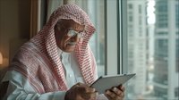 Senior Middle Eastern man in traditional red and white keffiyeh and thobe using digital tablet by window with urban skyline view in background.
