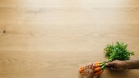 Fresh carrots with green leafy tops held in hand against wooden surface for cooking and healthy eating concepts.