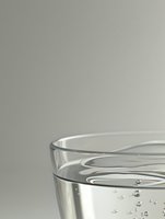 Clear glass of pure water with tiny air bubbles rising against neutral gray background, minimalist composition showing liquid refreshment and hydration.