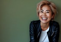 Confident mature Asian woman with short blonde hair wearing leather jacket and white t-shirt laughing genuinely against green background, expressing joy and authenticity.