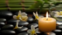 Zen spa composition with burning candle, white orchid flowers and black massage stones on bamboo mat for relaxation and wellness treatments.