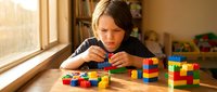 Young girl concentrating while building colorful block towers at wooden table by sunny window in cozy playroom setting for educational development.